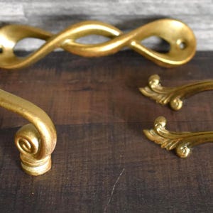 May include: A collection of antique gold-colored cabinet hardware, including two ornate handles with scroll details, a twisted handle, and a long, curved handle with decorative ends. The hardware is displayed on a dark wood surface.