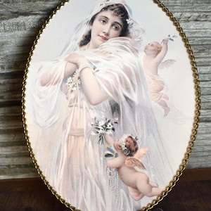 May include: Oval framed print featuring a woman in a white dress and veil, with two cherubs. The print has a gold-colored, beaded edge. The woman is holding flowers and a cherub is holding a branch.