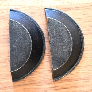 May include: Two matching, semi-circular, dark gray metal cabinet pulls. Each pull has a raised, curved outer edge and a textured, flat inner surface. The pulls are set against a light brown wood grain background.