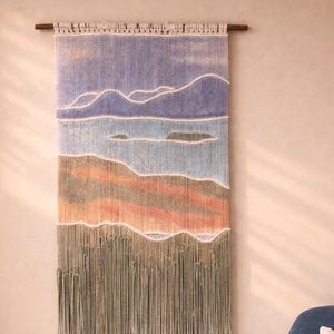 May include: A rectangular fiber wall hanging suspended from a dark wooden rod. The textile features a landscape design with horizontal bands of color including purple, light blue, tan, orange, and sage green, finished with long fringe.
