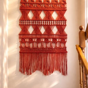 May include: A large, rectangular macrame wall hanging in a deep burnt orange color, suspended from a light-colored wooden dowel. The piece features intricate knot patterns, including geometric triangles, circular motifs, and a long, fringed bottom edge.
