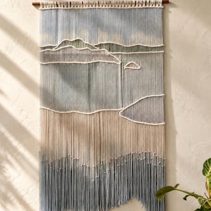 May include: A wall hanging made of cream and blue cotton cord, featuring a landscape design with layered mountain silhouettes created by thick horizontal cords. The piece is suspended from a wooden dowel, with long, loose fringe at the bottom.