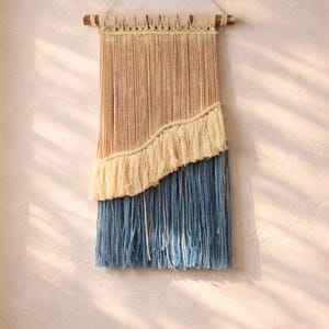 May include: A woven wall hanging with a natural beige top section, a cream-colored fringe, and a blue bottom section. The wall decor is suspended from a wooden dowel with a cream-colored cord.