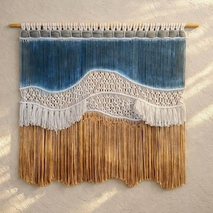 May include: A macrame wall hanging with a natural wood dowel. The design features a gradient of blue, white, and tan yarn, creating a layered, textured effect. The top section is gray, transitioning to blue, with a white, woven middle and tan fringe.