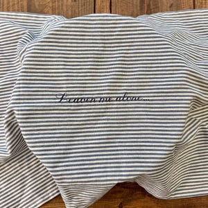 May include: A folded, striped fabric with alternating white and blue-grey lines. The phrase "Leave me alone..." is embroidered in a cursive font. The fabric is resting on a wooden surface.