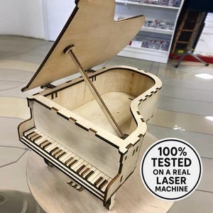 以下が含まれることがあります： 木製のピアノ型の箱で、蓋が開いており、木製の支柱で支えられています。 キーボードには黒と白のキーがあります。 「100％ TESTED ON A REAL LASER MACHINE」というテキストが丸いステッカーにあります。