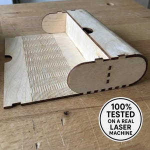 May include: A wooden box with a curved lid and base, featuring intricate laser-cut details. The box is made of light-colored wood and has a rectangular shape with a curved top. The image includes a circular sticker that says "100% TESTED ON A REAL LASER MACHINE."