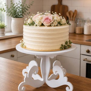 Può includere: Una torta rotonda con una liscia glassa color bianco sporco, guarnita con un bouquet di rose rosa, fiori bianchi e foglie verdi. La torta poggia su un'alzata bianca con gambe decorative curve e una piattaforma circolare.