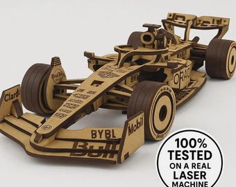 May include: A wooden model of a Formula 1 race car. The car is constructed from light brown wood with dark brown wheels and accents. The model features detailed cutouts and sponsor logos. A circular sticker on the side reads "100% TESTED ON A REAL LASER MACHINE."