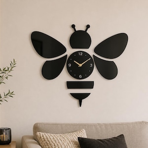 May include: A wall clock shaped like a bee, featuring a black circular clock face with gold hands and numerals at 3, 6, 9, and 12. The bee design includes black wings, a head with antennae, and a segmented body, all mounted as separate pieces.