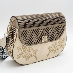 May include: Wooden handbag with a laser-cut design. The bag features a geometric pattern on the top and a floral design on the bottom. It has a beige strap with a black and white geometric pattern and gold-colored hardware.