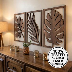 May include: Three wooden wall art panels with leaf designs. The panels are framed in dark brown wood and feature intricate cut-out patterns of leaves. The panels are mounted on a light-coloured wall above a wooden cabinet. The image also includes a vase and candle holders.