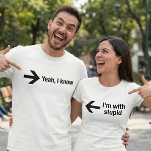May include: Two people wearing white t-shirts with black text and arrows. One shirt says "Yeah, I know" with an arrow pointing right. The other says "I'm with stupid" with an arrow pointing left. They are smiling and pointing at the text.