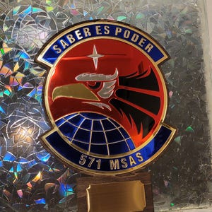 May include: A military emblem with the text "SABER ES PODER" in a blue and red design. The emblem features an eagle and the text "571 MSAS". It is mounted on a wooden base with a gold plaque.