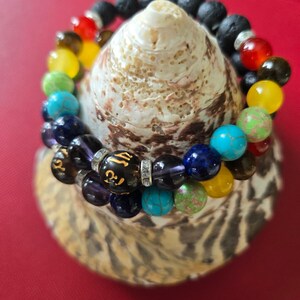 May include: Two beaded bracelets with colorful gemstones and black lava beads are displayed on a seashell. The bracelets feature a variety of colors, including red, orange, yellow, green, blue, and purple. The bracelets are resting on a red surface.