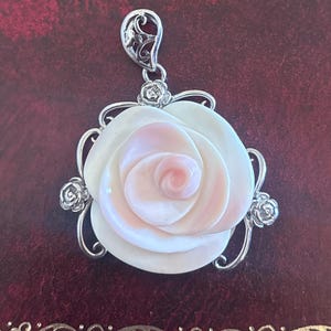 May include: A pendant featuring a carved rose design. The rose is white with a pink center, set in a silver-toned filigree frame. The pendant has a decorative bail and small rose accents.