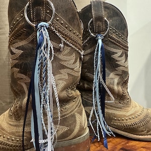 May include: A pair of distressed brown leather cowboy boots featuring decorative silver-toned metal rings attached to the pull tabs. Each ring is adorned with a cascading arrangement of navy blue, light blue, and silver ribbons and chains.