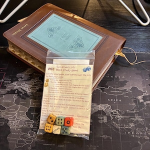 May include: A brown leather-bound Holy Bible with a light blue rectangular inlay on the cover, positioned behind a clear plastic bag containing a Scripture Lots Bible study game with six small wooden dice in yellow, blue, and orange colors.