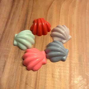 May include: A collection of six seashell-shaped objects in various colors, including red, pink, light blue, lavender, and gray. The shells are arranged on a wooden surface, showcasing their glossy finish and detailed ridges.