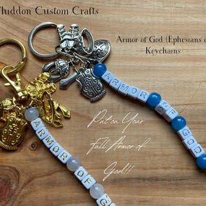 May include: Two keychains featuring silver and gold-toned metal charms shaped like helmets, shields, and swords. Each keychain includes a string of white letter beads spelling ARMOR OF GOD, accented with blue or white round beads on metal rings.