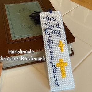 May include: Handmade Christian bookmark with the text "The Lord is my Shepherd" in blue thread. The white bookmark features two yellow crosses and a blue tassel. The bookmark is next to a brown book with a blue cover.
