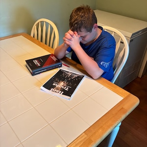 May include: A person wearing a blue short-sleeved shirt with hands clasped in prayer. On the table are a black and red Foundation Study Bible and a white booklet titled Hard Truths with a black and white graphic image on the cover.