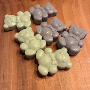 May include: A collection of small, bear-shaped objects in shades of pale green and lavender. The objects are arranged on a wooden surface, with the bears' details clearly visible. The lighting highlights the smooth texture of the bears.