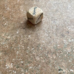 May include: A small, beige, cube-shaped object with handwritten black text on each side. The text includes the words "Proverbs", "Genesis", and "Ephesus". The cube is resting on a mottled brown surface.