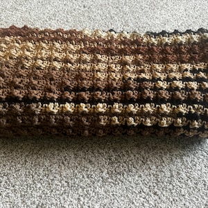 May include: A crocheted scarf in shades of brown, tan, and black. The scarf has a textured, ridged pattern and is folded over. The yarn colors create horizontal stripes across the scarf. The scarf is lying on a gray surface.