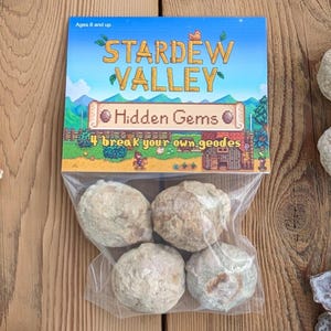 May include: A clear plastic bag containing four geodes, with a Stardew Valley Hidden Gems package on top. The package features the game's logo and the text "4 break your own geodes". The background is a wooden surface.
