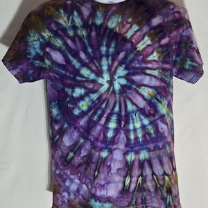 Può includere: Una maglietta tie-dye a maniche corte con un motivo a spirale. La maglietta presenta una miscela di colori, tra cui sfumature di viola, blu e verde. Il design si irradia dal centro, creando un effetto visivamente sorprendente. La maglietta è esposta su un manichino.