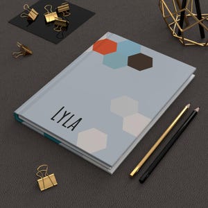 May include: A light blue journal with a geometric design of orange, blue, and brown hexagons. The name "LYLA" is printed in black on the cover. Gold and black pencils and gold binder clips are also visible.