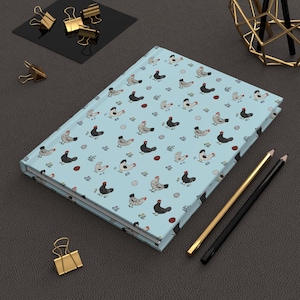 May include: A light blue journal with a repeating pattern of chickens, eggs, and sprigs. The journal is lying on a dark gray surface, with gold and black pencils, and gold binder clips nearby.