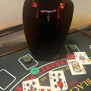 May include: A black beaded necklace with red and black accents, featuring a silver number 7 pendant and red cube-shaped beads. The necklace is displayed on a black bust, with playing cards and casino chips in the background.
