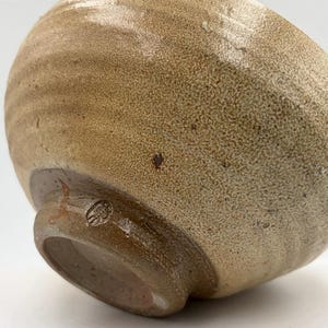 Puede incluir: Un cuenco de cerámica hecho a mano con un exterior marrón claro y texturizado. El cuenco tiene una forma redondeada con una pequeña base circular y una marca estampada. El borde es de un tono marrón más oscuro.