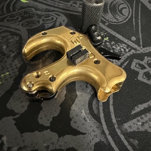 May include: A gold-colored archery release aid with a curved handle and a black trigger. The release aid has a textured, cylindrical grip and two black pads. The device is designed for use with a bow and arrow.