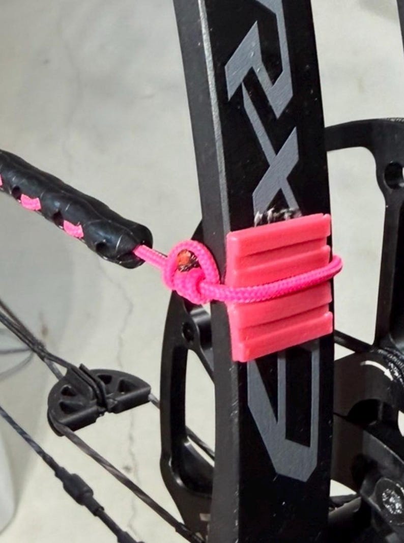 Peut inclure: Gros plan sur un arc &agrave; poulies noir avec une corde rose et un repose-fl&egrave;che rose. L'arc porte les lettres "RX" en argent. La corde rose est nou&eacute;e autour du cadre de l'arc.