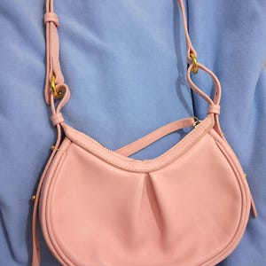 May include: A light pink shoulder bag with a curved shape and a long, adjustable strap. The bag has a zipper closure and gold-colored hardware. The bag is made of a smooth material and has a simple, elegant design