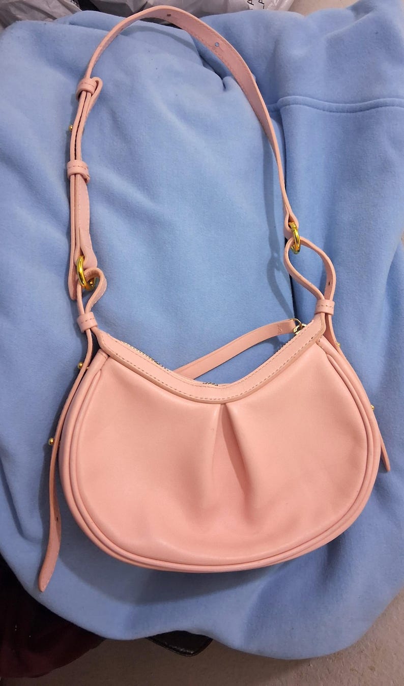 May include: A light pink shoulder bag with a curved shape and a long, adjustable strap. The bag has a zipper closure and gold-colored hardware. The bag is made of a smooth material and has a simple, elegant design