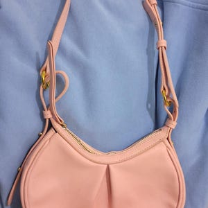 May include: A light pink shoulder bag with a curved shape and a gold zipper. The bag has a long, adjustable strap and gold-colored hardware. The bag is made of a smooth material and has a small logo on the front