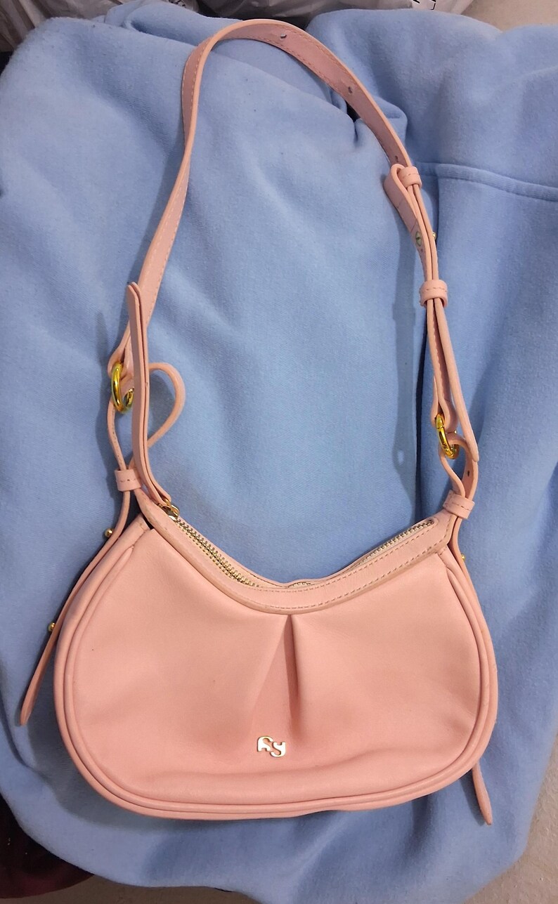 May include: A light pink shoulder bag with a curved shape and a gold zipper. The bag has a long, adjustable strap and gold-colored hardware. The bag is made of a smooth material and has a small logo on the front