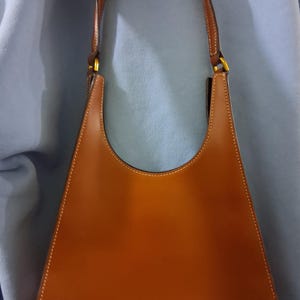 以下が含まれることがあります： 曲線的な上部と三角形の形をした茶色の革製ハンドバッグ。バッグは茶色のハンドルと金色の金具が付いています。白いステッチがバッグの端を強調しています。
