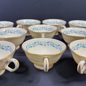 Könnte beinhalten: Ein Satz cremefarbener Teetassen mit goldenen Verzierungen und Henkeln. Jede Tasse zeigt ein zartes blaues und gelbes Blumenmuster am Rand. Die Tassen sind auf einer dunklen Oberfläche angeordnet und präsentieren ihr elegantes Design.