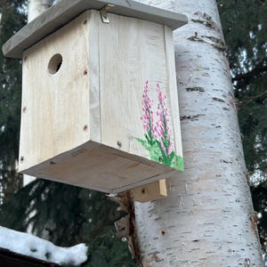 May include: A wooden birdhouse with a grey roof is mounted on a tree. The birdhouse features a floral design in pink and green. It has a circular entrance hole and a small perch. The birdhouse is attached to a tree.