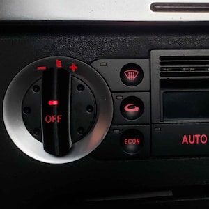 Puede incluir: Primer plano del panel de control de clima negro de un coche. Cuenta con diales, botones y una pequeña pantalla. La palabra "AUTO" se ilumina en rojo, junto con otros símbolos e indicadores.