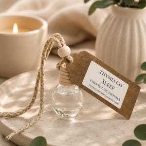 May include: A small clear glass essential oil diffuser bottle with a wooden cap and a braided beige cord featuring a light wooden bead. A brown paper tag is attached, printed with the text THYMELESS SLEEP ESSENTIAL OIL DIFFUSER THYMELESSCOMYSHOPIFY.COM.