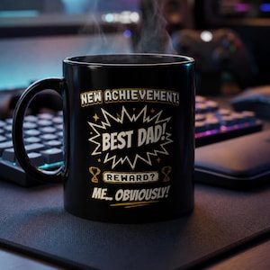 May include: Black coffee mug with the text "NEW ACHIEVEMENT! BEST DAD! REWARD? ME...OBVIOUSLY!" in white and gold lettering. The mug is on a desk with a keyboard and mouse.