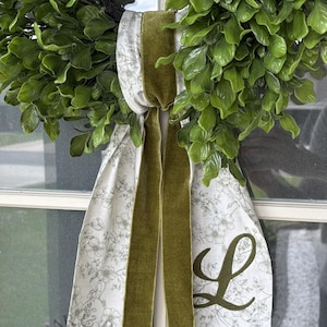 May include: A green boxwood wreath adorned with a hanging fabric ribbon featuring a white and grey floral pattern. The ribbon includes a central olive green velvet strip and a large, dark green embroidered letter L on the right side.