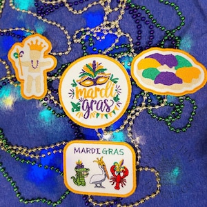 May include: Embroidered Mardi Gras patches in various shapes, including a baby, a mask, a king cake, and a rectangular patch with an alligator, pelican, and crawfish. The patches are white with yellow, green, purple, and orange accents, and are surrounded by beaded necklaces.