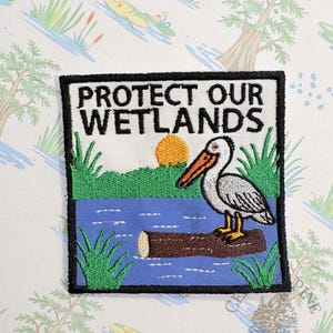 May include: Embroidered patch with the text "PROTECT OUR WETLANDS". The patch features a pelican on a log in a blue body of water, with green grass and a yellow sun. The patch has a black border.